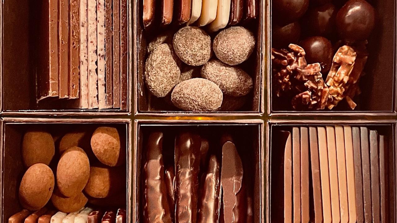 A close-up of an assorted chocolate box divided into six compartments, each holding multiple different types of chocolates: truffles, chocolate-dipped sticks, caramel clusters, and layered bars.
