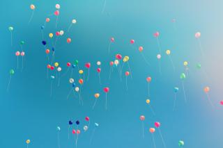 Dozens of small, brightly coloured balloons drifting upward against a clear blue sky.