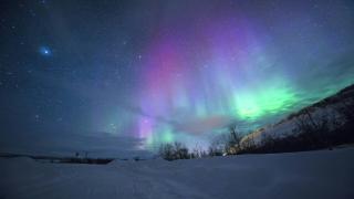 A vivid green and pink aurora arcing across a star-filled night sky over a snow-covered landscape.
