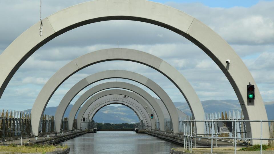 Repeated concrete arches of the Falkirk Wheel aqueduct receding in perspective over a still canal channel, with a green canal-traffic signal on the right and distant hills visible through the openings under broken cloud.
