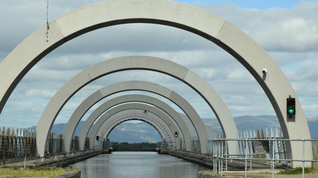 Repeated concrete arches of the Falkirk Wheel aqueduct receding in perspective over a still canal channel, with a green canal-traffic signal on the right and distant hills visible through the openings under broken cloud.
